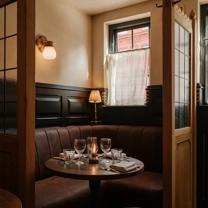 A cozy restaurant booth with a round table set for four, featuring wine glasses, plates, and a candle. The booth has brown leather seating, soft lighting, and a window with sheer curtains.