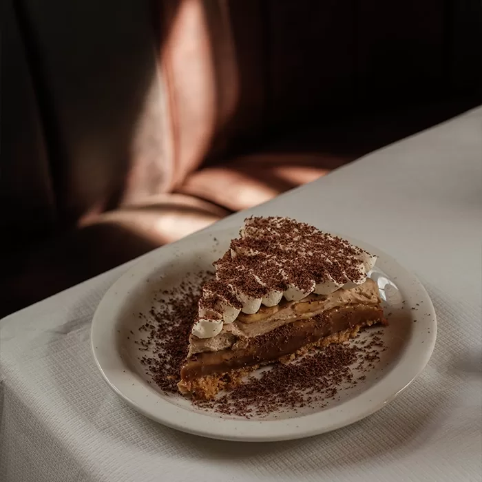 A slice of creamy pie topped with whipped cream and chocolate shavings sits on a white plate, with extra chocolate sprinkled around, on a white tablecloth in a softly lit, cozy setting.