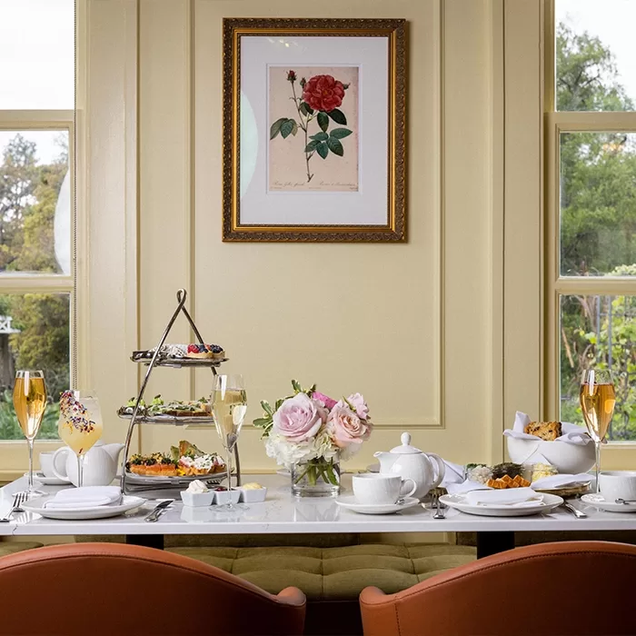 A table set for afternoon tea with teapots, cups, pastries, sandwiches, scones, and champagne flutes. A floral centerpiece and tiered tray are on the table; a framed rose illustration hangs on the cream wall behind.
