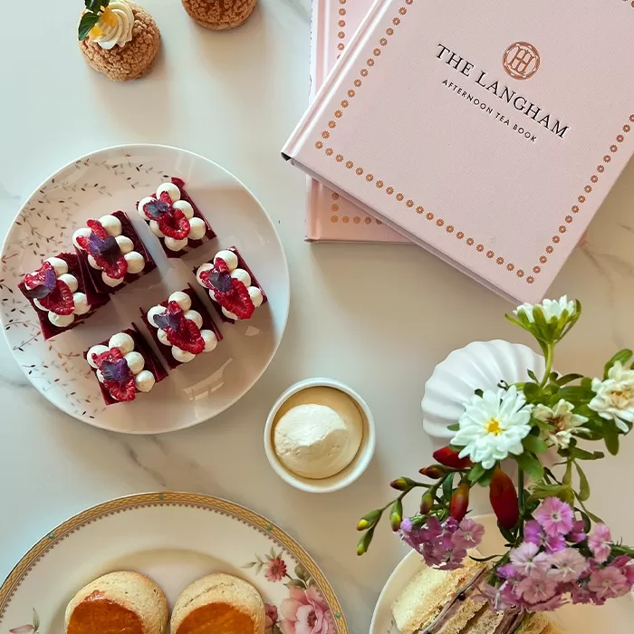 A table set with elegant pastries, scones, a dish of cream, a floral arrangement, and two pink The Langham Afternoon Tea Book boxes on a white surface.