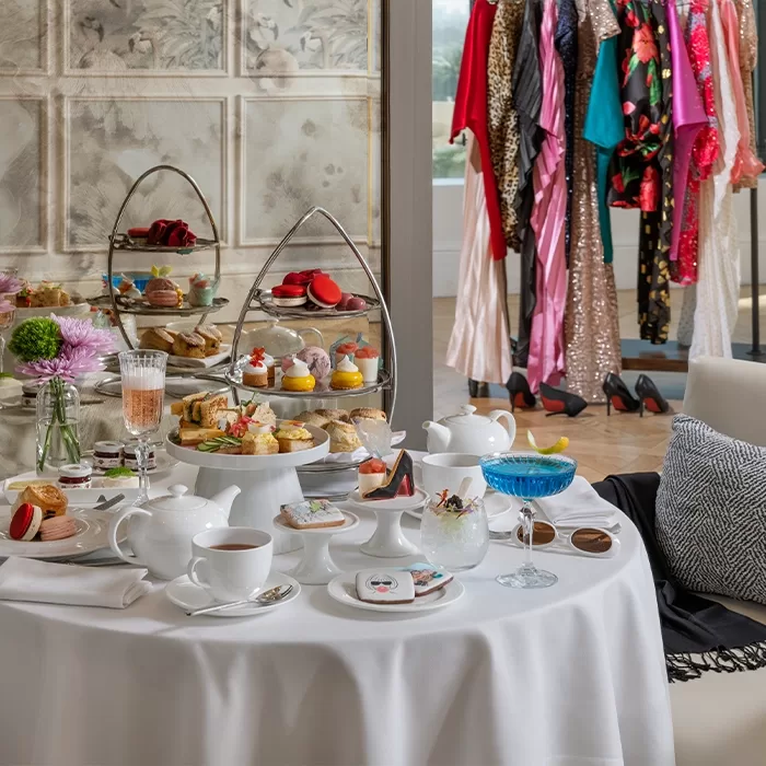 An elegant afternoon tea setup with teapots, cups, pastries, and desserts on a round table, with tiered serving trays. In the background, a clothing rack displays colorful and sparkling dresses.
