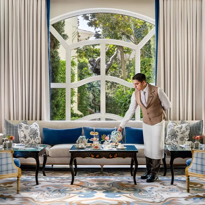 A waiter in a beige vest and white apron serves tea in an elegant, sunlit sitting room with a large window, plush sofa, patterned rug, and trees visible outside.