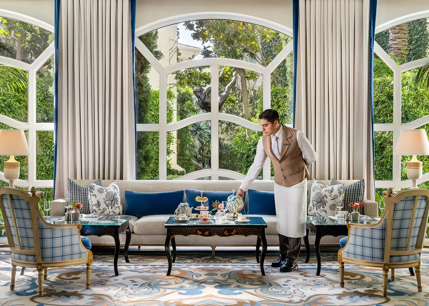 A waiter in a vest and apron serves afternoon tea on a coffee table set with teapots, cups, and tiered trays in an elegant, sunlit lounge with large arched windows, classic furniture, and garden views.