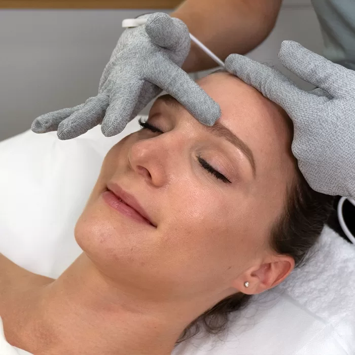 A woman lies on a treatment bed with her eyes closed while someone wearing gray gloves gently massages her forehead, performing a facial treatment.