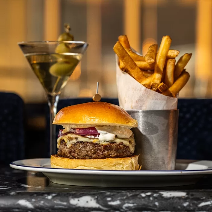 A cheeseburger with onions and sauce on a bun sits on a plate next to a metal cup of French fries. In the background, there is a martini with green olives on a pick.