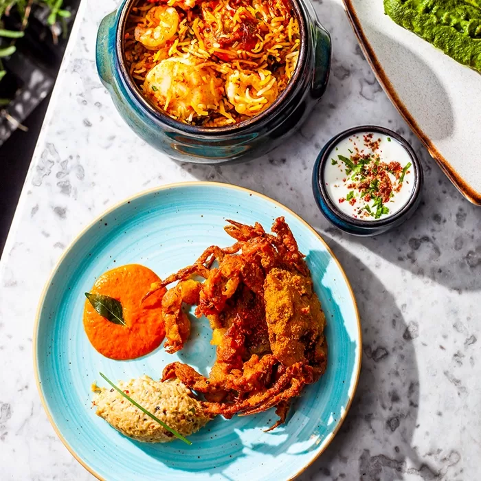 A blue plate with fried soft-shell crab, a dollop of beige dip, and a smear of orange sauce. Nearby, a bowl of biryani with shrimp and egg, and a small bowl of yogurt with herbs, all on a marble table.
