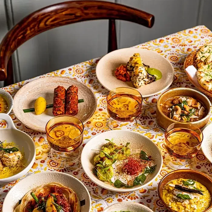 A table set with various Indian dishes in beige bowls, including curries, kebabs, naan, and colorful garnishes. Amber glasses hold drinks, and the tablecloth features a yellow floral pattern. A wooden chair is partially visible.