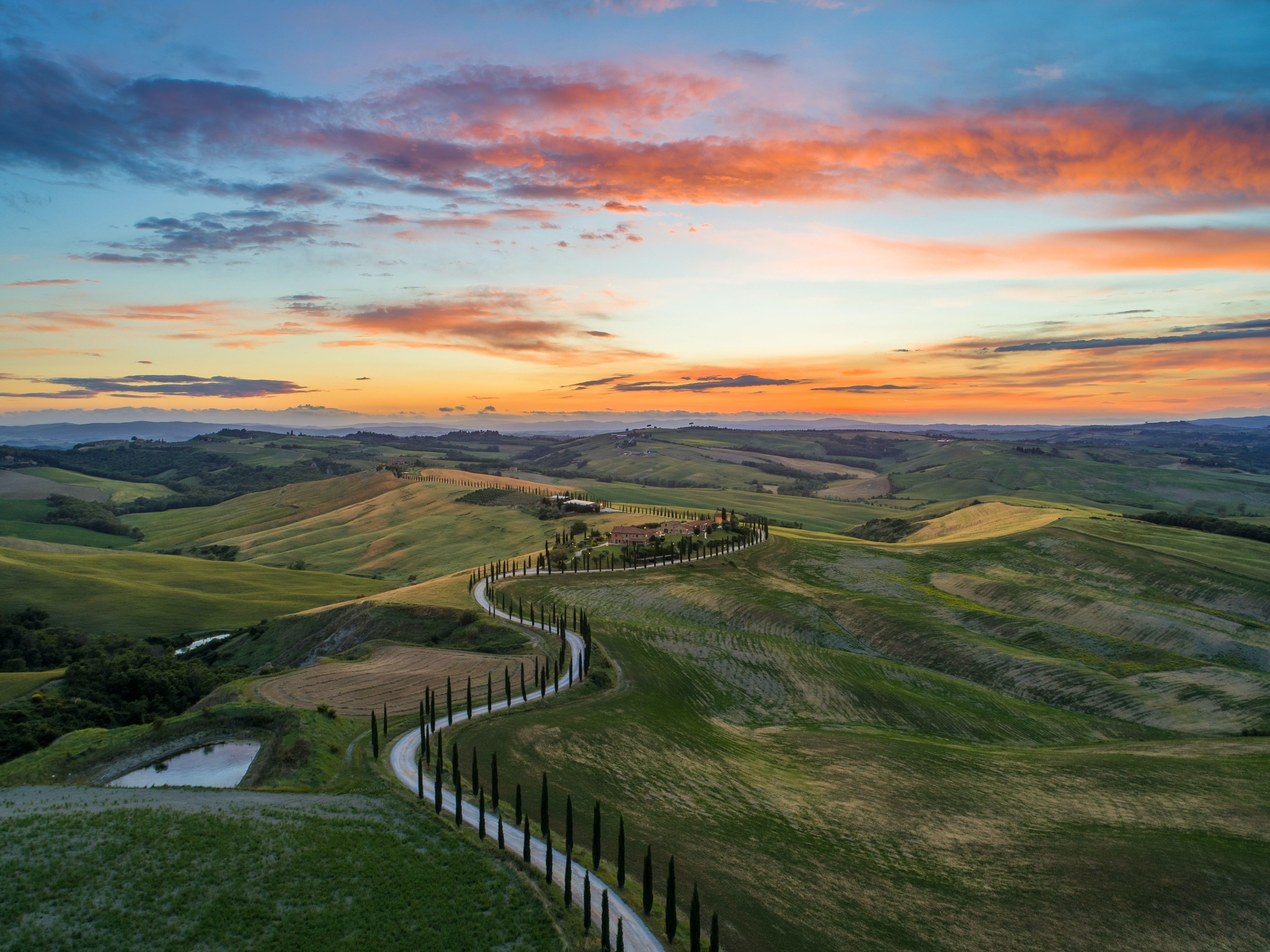 Birdseye view of Tuscan countryside hills with long winding path with rows of little trees guiding the path either side with orange and red cloudy sky at sunset