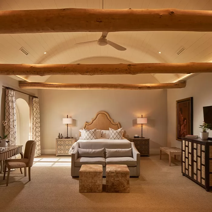 A spacious, warmly lit bedroom with exposed wooden ceiling beams, a large bed with a decorative headboard, matching nightstands, lamps, a bench, woven stools, and a seating area near arched windows with patterned curtains.