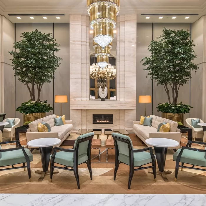 Elegant hotel lobby with tall trees, modern chandeliers, beige sofas, green armchairs, a marble fireplace, round tables, and soft lighting, creating a sophisticated and inviting atmosphere.