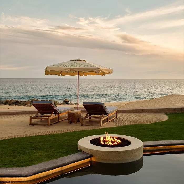 Two wooden lounge chairs and a beige umbrella sit on a sandy beach facing the ocean at sunset, with a round fire pit in the foreground next to a pool and green grass.