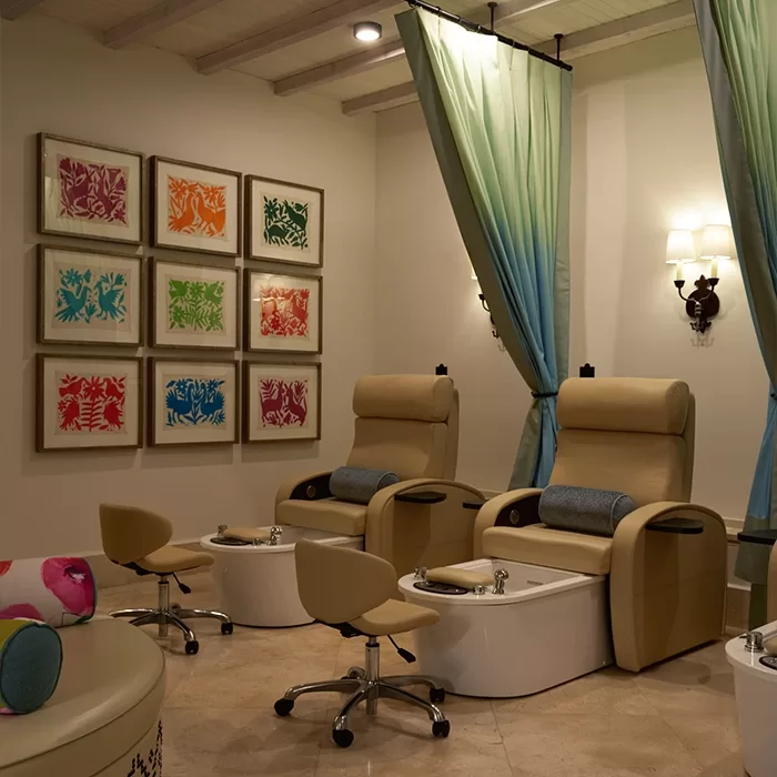 Two beige pedicure chairs with foot baths and rolling stools sit in a cozy spa room. Colorful framed art hangs on the wall, and soft green curtains divide the space. Warm lighting adds to the relaxing atmosphere.