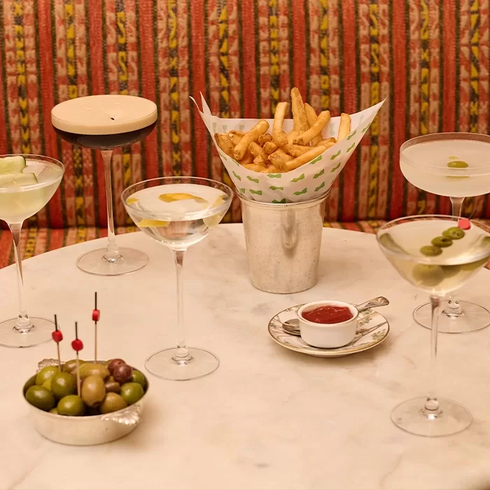 A round table with four martinis, a coupe glass with a frothy drink, a metal cup of French fries, a small bowl of ketchup, and a dish of green and black olives on toothpicks, set against a patterned red background.