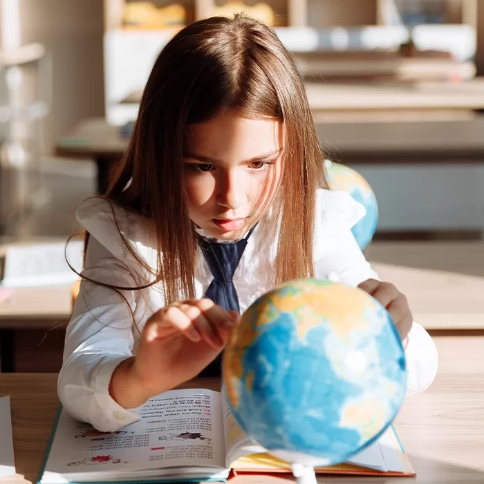 A young girl with long brown hair, wearing a white shirt and tie, sits at a desk studying a globe with a book open in front of her in a brightly lit classroom.