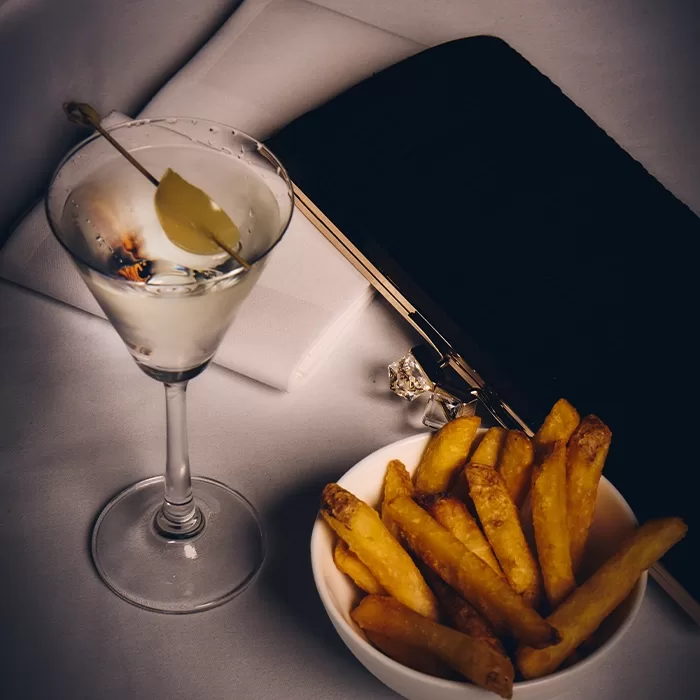 A martini with an olive, a bowl of French fries, a black clutch bag, and a folded white napkin on a table with a neutral background.