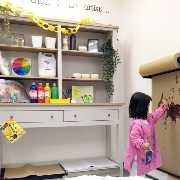 A young child in a pink smock paints on a large sheet of brown paper attached to a wall. Behind her is a shelf with art supplies, plants, and decorations in a well-organized, creative workspace.