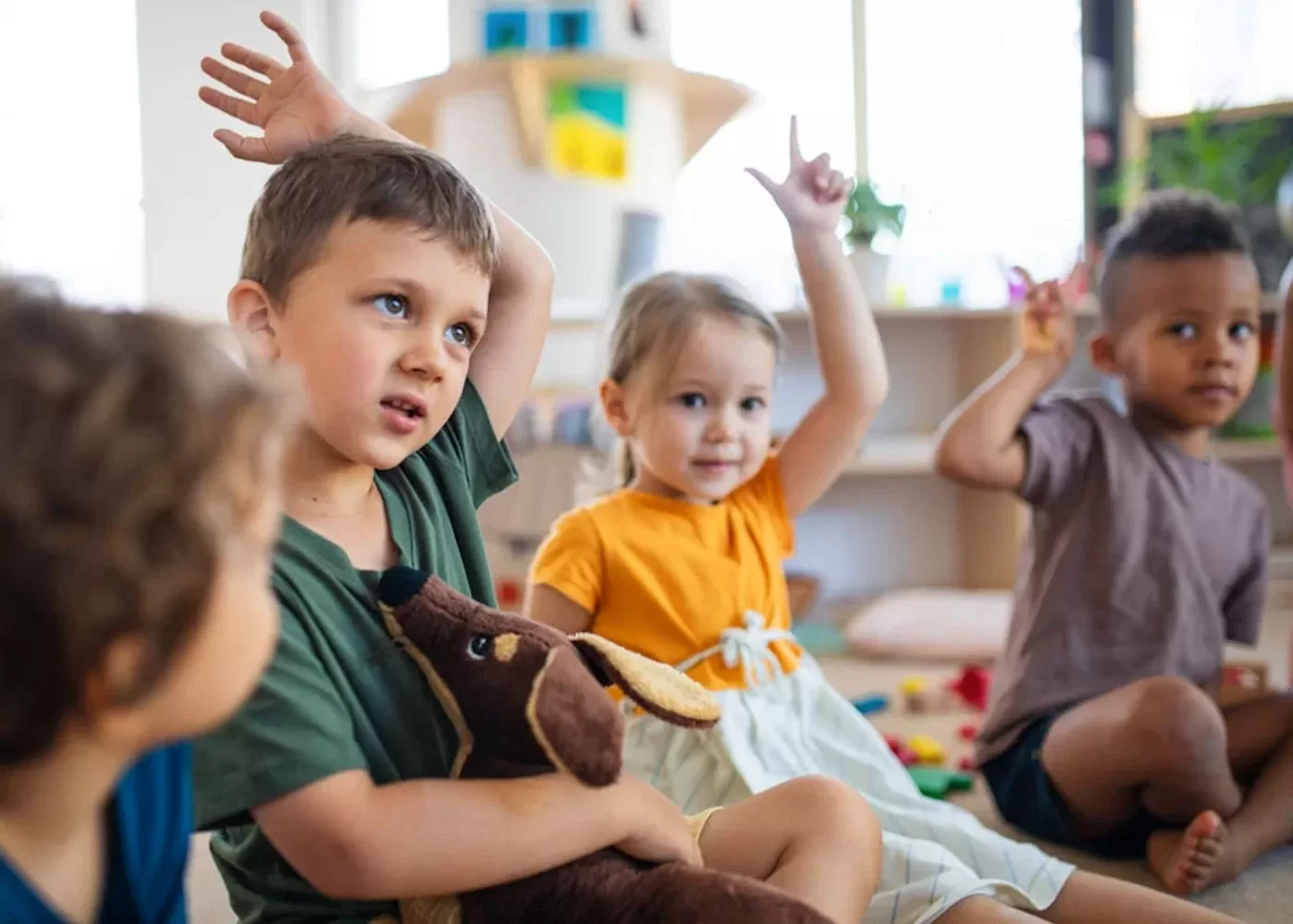Four young children sit on the floor in a classroom, raising their hands eagerly. One boy in front holds a plush dog toy. The children look attentive and engaged, with shelves and toys visible in the background.