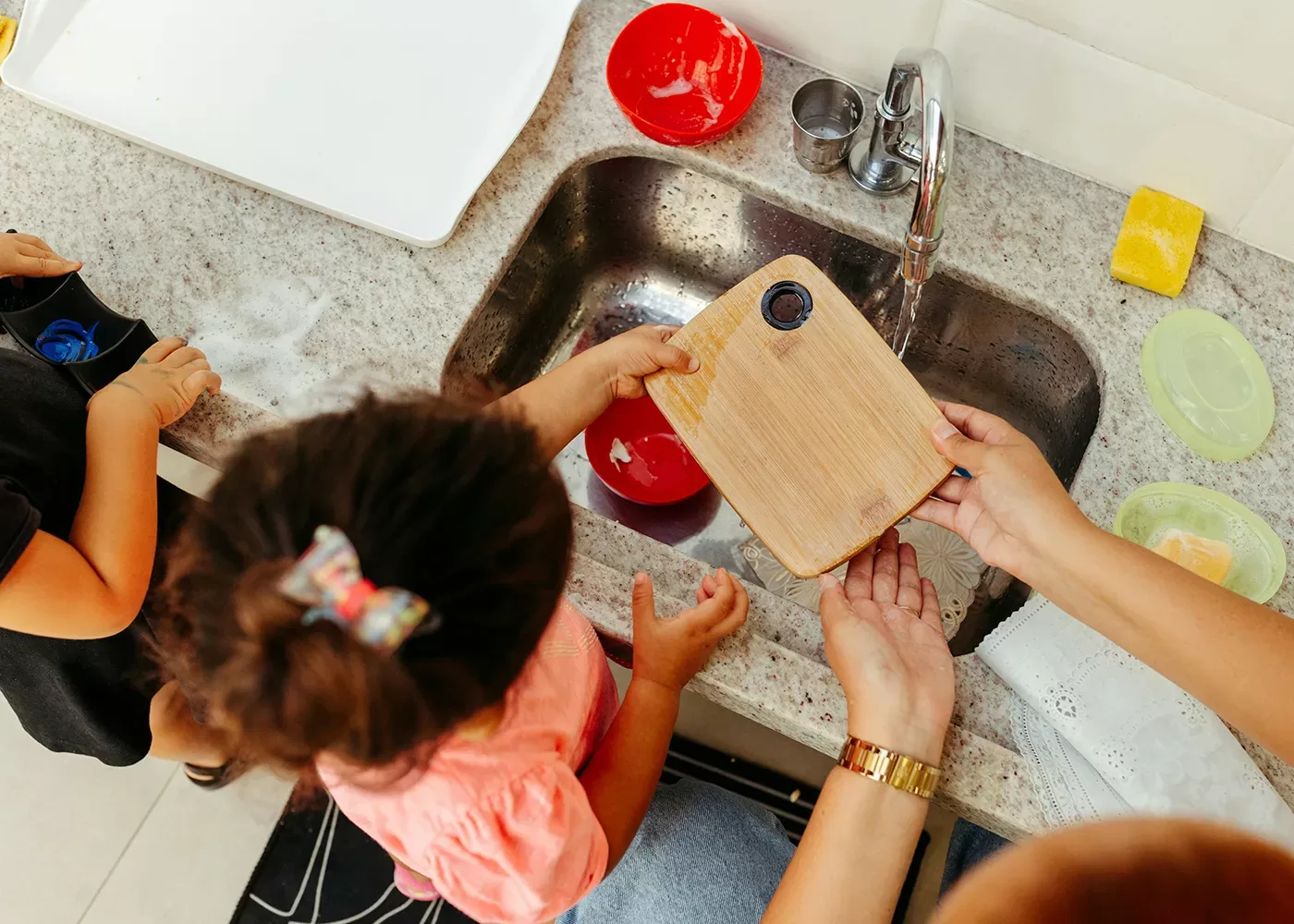 A child and an adult are washing a small wooden cutting board in a kitchen sink. The countertop is cluttered with soap, sponges, and a red bowl. The child is wearing a pink shirt, and the adult is wearing a watch on the left wrist.