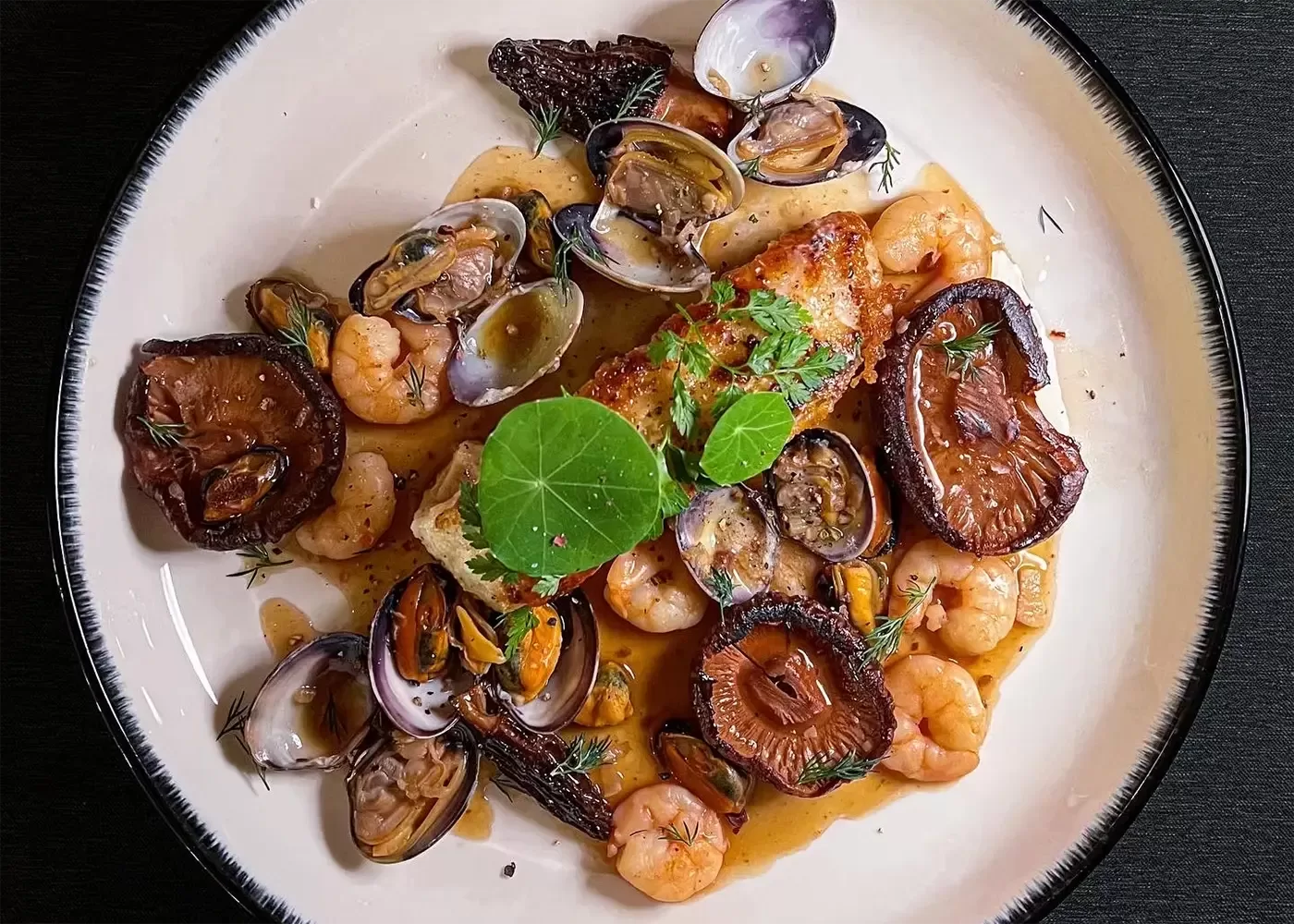A gourmet dish featuring seared fish topped with herbs, surrounded by shrimp, clams, mushrooms, and a rich brown sauce, artfully arranged on a white plate with a dark rim.