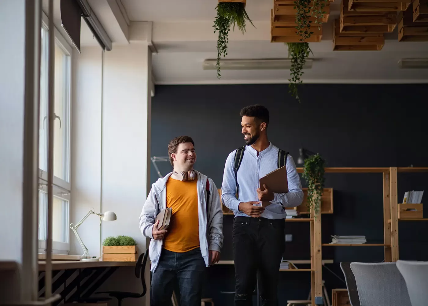 Two men walking indoors, smiling and carrying folders. The one on the left wears a gray hoodie and yellow shirt, with headphones around his neck. The one on the right wears a light blue shirt and dark pants. Theyre in a modern office with plants and shelves.