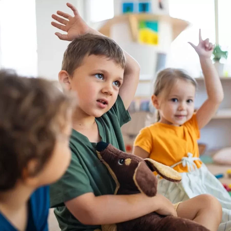 Three young children sit indoors; one boy in front holds a stuffed dog and has his hand raised thoughtfully, while a girl in an orange shirt and another boy nearby also raise their hands, likely in a classroom setting.