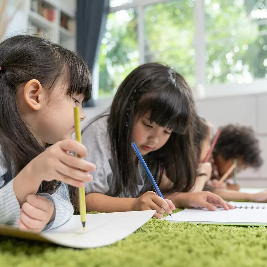 Four young children lie on the floor inside, drawing or writing in notebooks with colored pencils. They are focused on their work, surrounded by natural light from large windows in the background.
