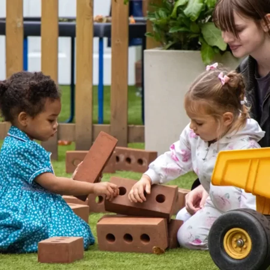 Two young girls play with toy bricks on grass while a woman sits nearby, watching and smiling. A toy yellow dump truck is beside them, and a wooden fence and greenery are in the background.