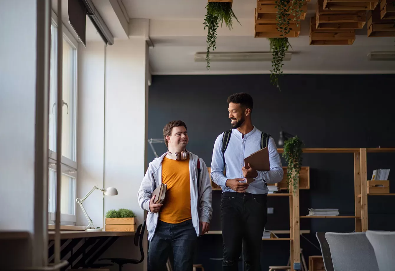 Two men walk through an office with large windows. One wears an orange shirt and has headphones, while the other wears a white shirt and carries a laptop. They are smiling and appear to be in conversation. The office has plants and modern decor.