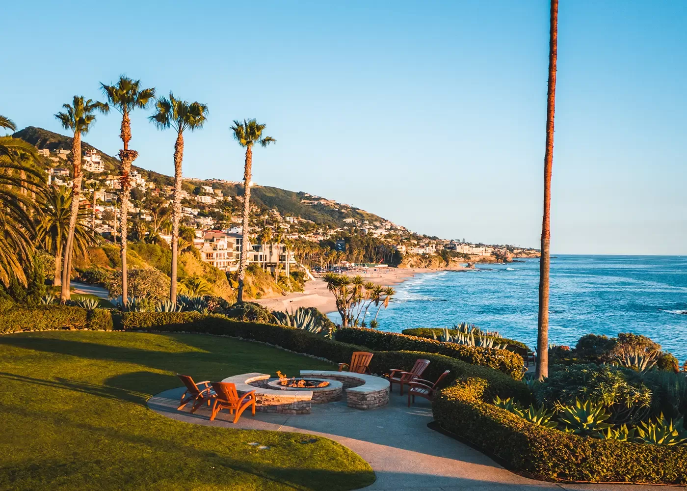 Sunny coastal view with palm trees, a fire pit area surrounded by chairs, manicured grass, and ocean waves. Hills with houses overlook the sandy beach and blue sea under clear skies.