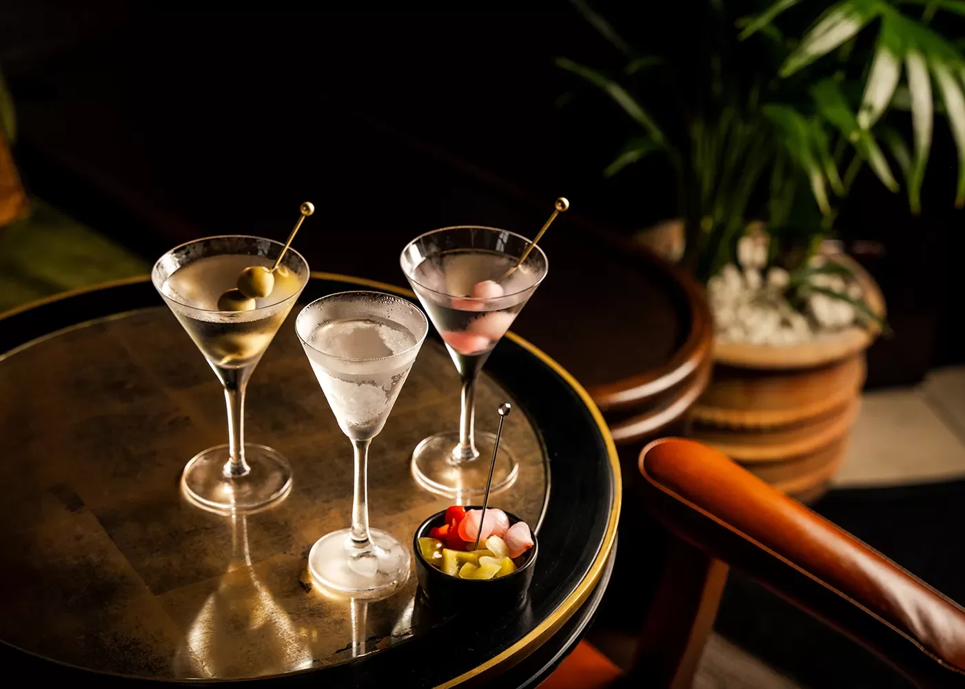 Three martini glasses with olives and cocktail onions are arranged on a round, reflective table next to a small bowl of garnishes. There is an orange chair and a potted plant in the background.