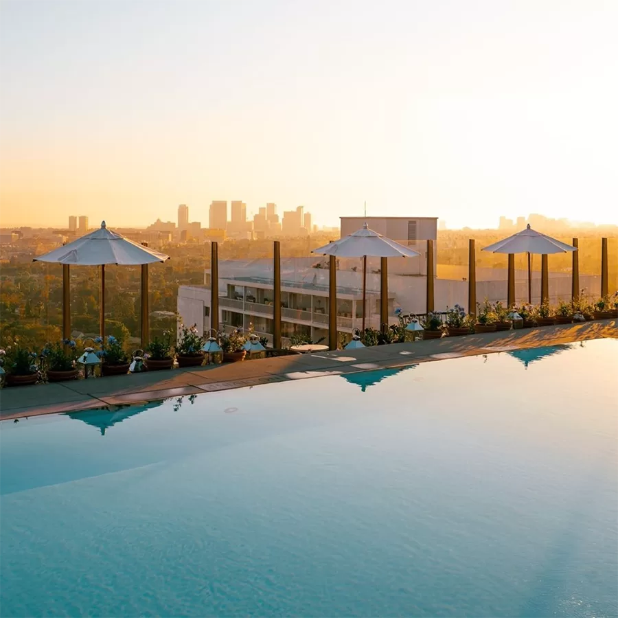 A rooftop swimming pool with white umbrellas and flower planters overlooks a city skyline at sunset, with tall buildings visible in the distance under a clear sky.