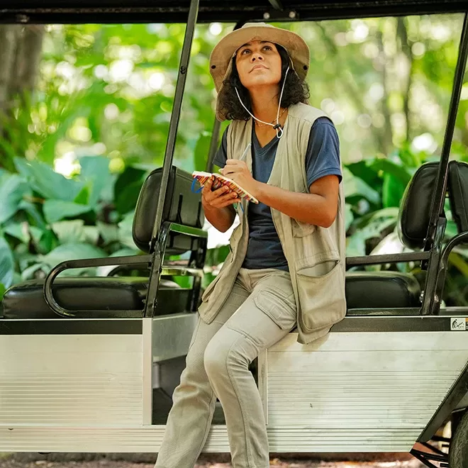 A person in outdoor clothing and a hat sits on the edge of a vehicle, holding a notebook and looking upward, surrounded by green foliage in a forest or jungle setting.