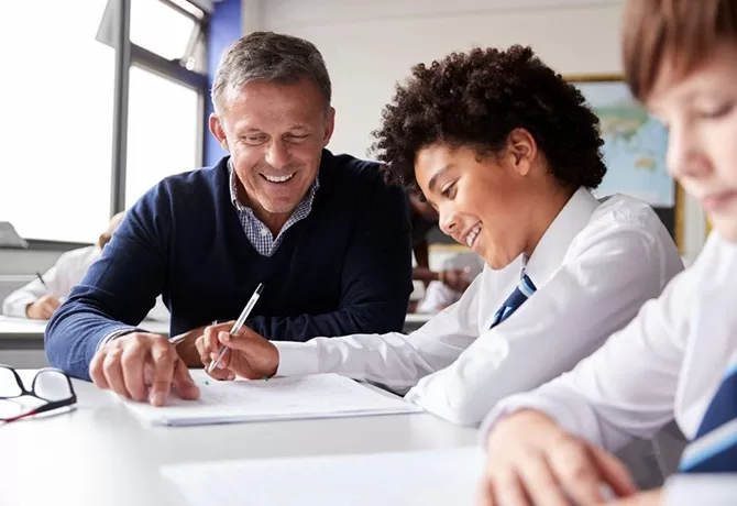 Male teacher and students smiling, sitting at desk in a school completing work with a pen and paper.