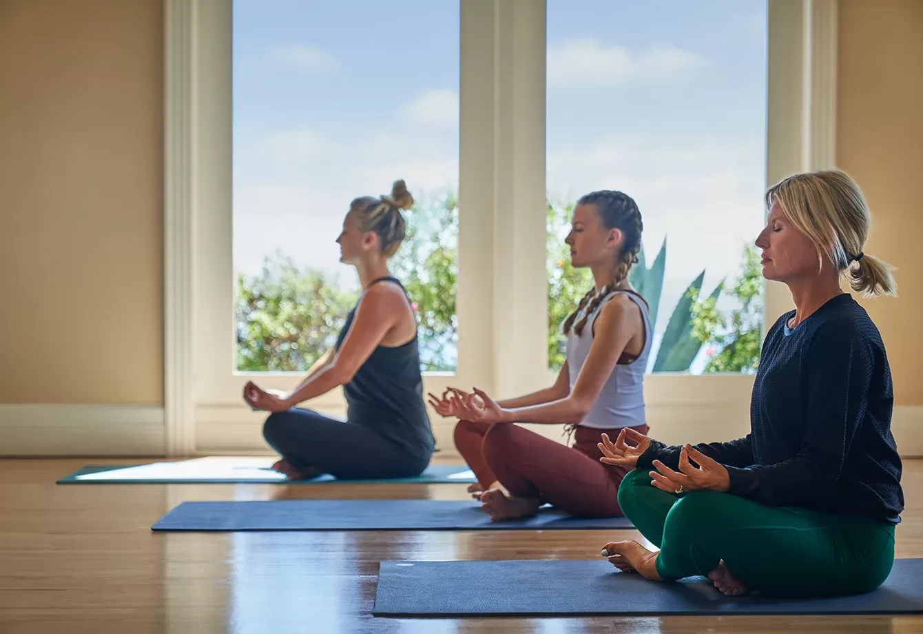 Three women sit cross-legged on yoga mats indoors, meditating with eyes closed and hands resting on knees. Large windows behind them reveal green plants and a bright, peaceful outdoor scene.