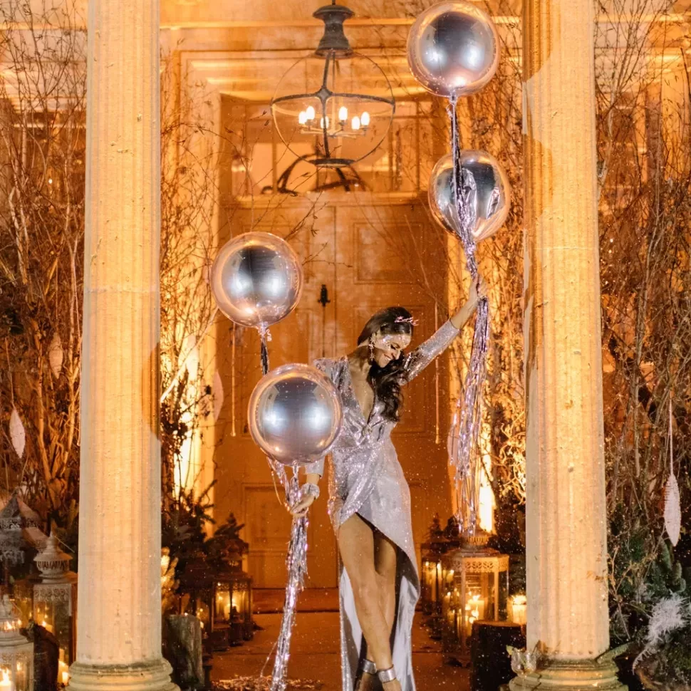 A woman in a shimmering silver dress gracefully holds a cluster of silver balloons, creating an elegant scene.