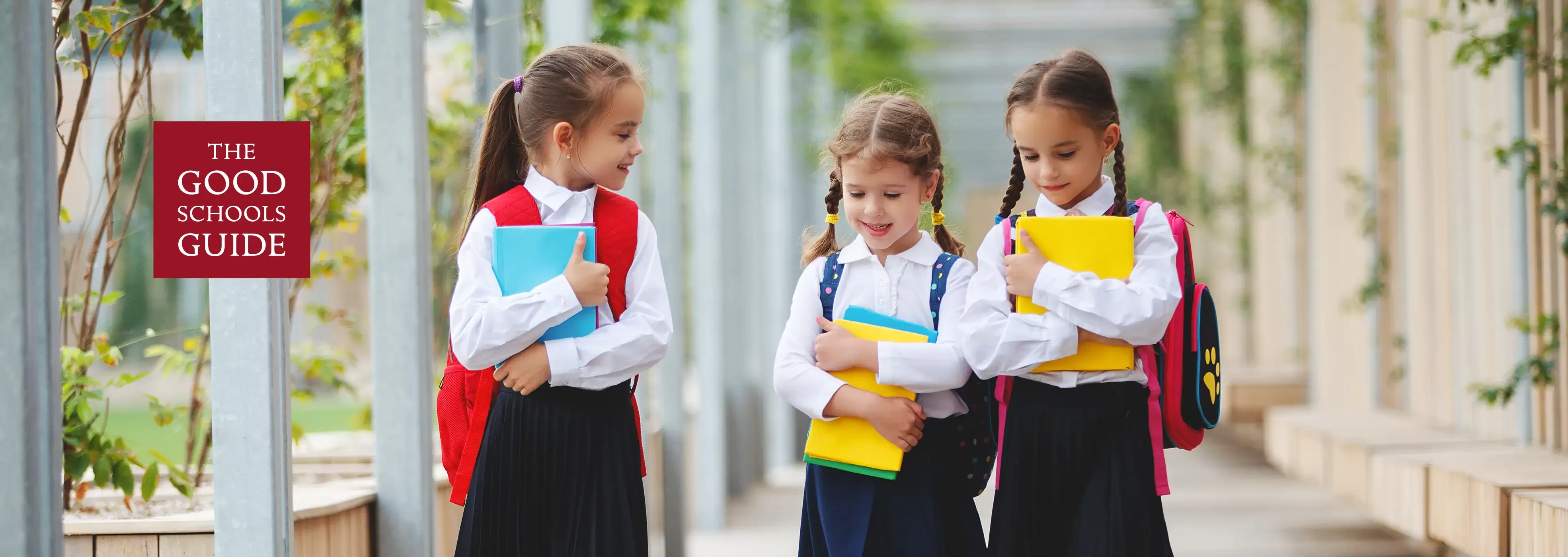 Three young schoolgirls in uniform walk outdoors, smiling and carrying colorful books and backpacks. A sign on the left reads The Good Schools Guide. The background features greenery and columns.