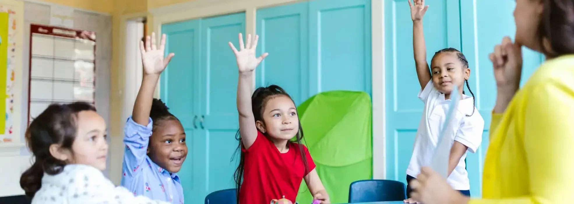 Four young children in a classroom sit at a table, three with hands raised, smiling and looking toward a teacher standing in front of them. Blue cabinets and a green board are visible in the background.