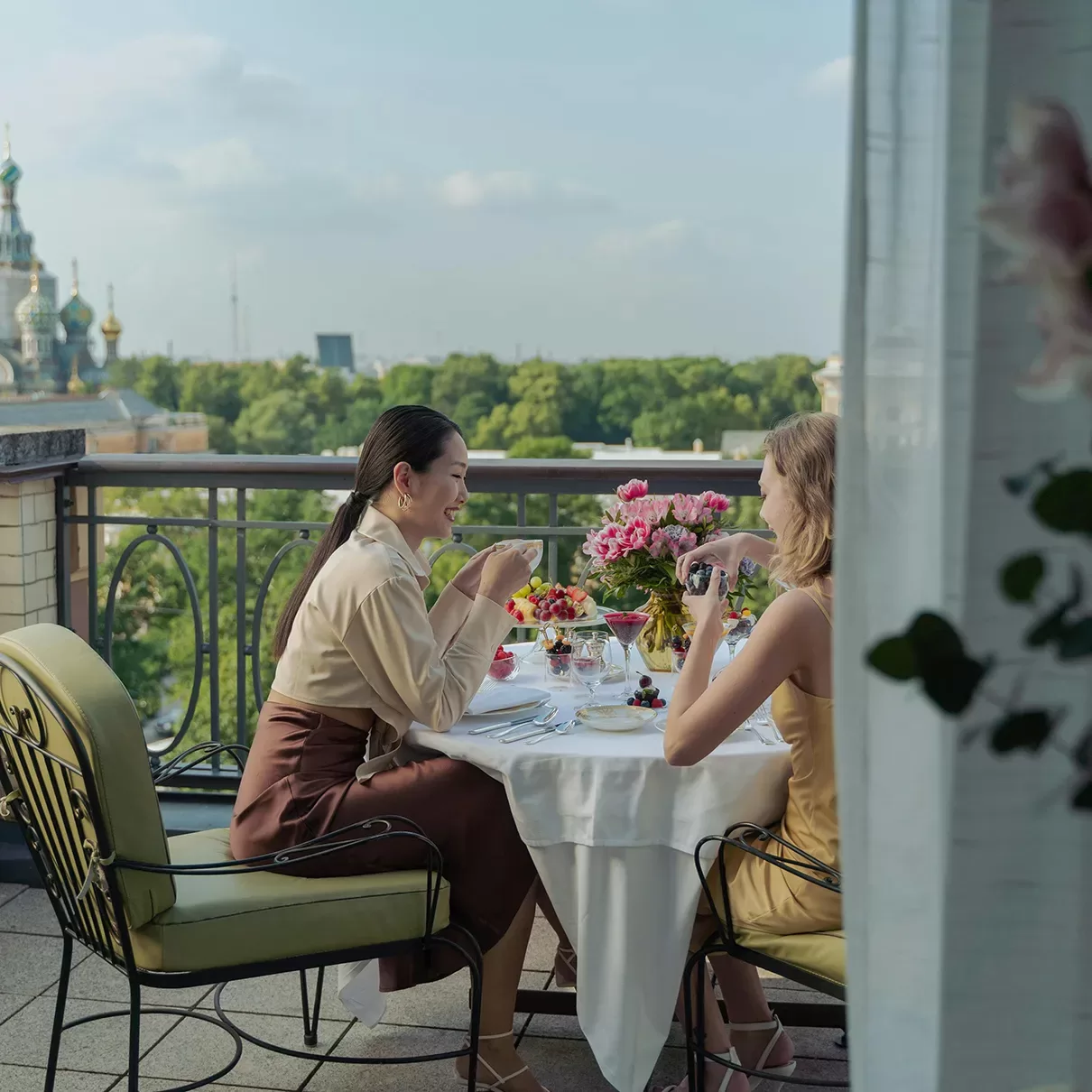Two women sit at an outdoor table on a balcony, enjoying drinks and desserts. The table is set with flowers and glassware. In the background, trees and a distant ornate building are visible under a blue sky.